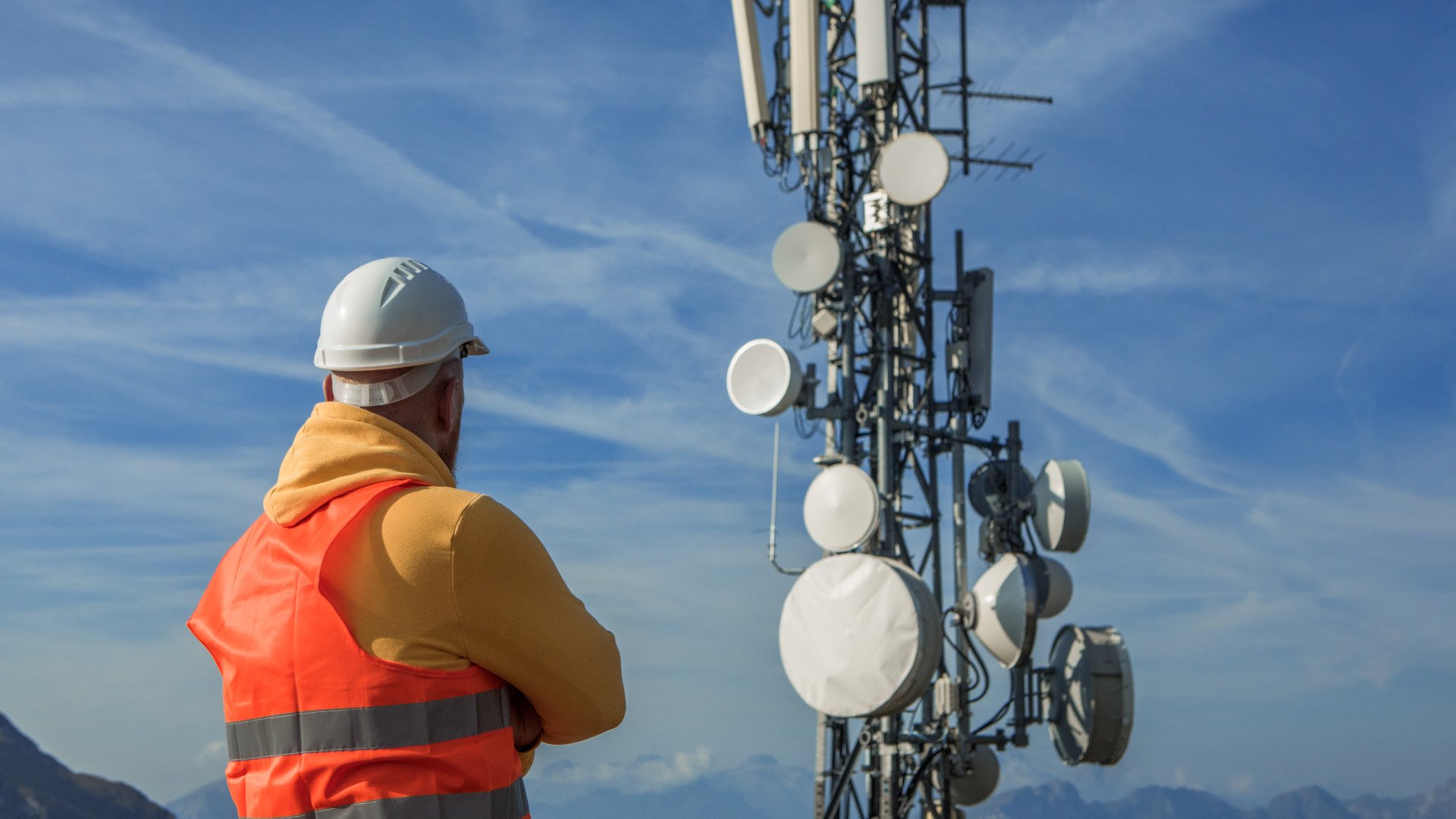 Ein Techniker mit Helm und Warnweste steht vor einem Mobilfunkmast in malerischer Landschaft